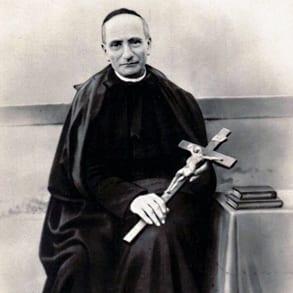 Black-and-white photograph of a priest in cassock seated and holding a large crucifix, with books nearby.